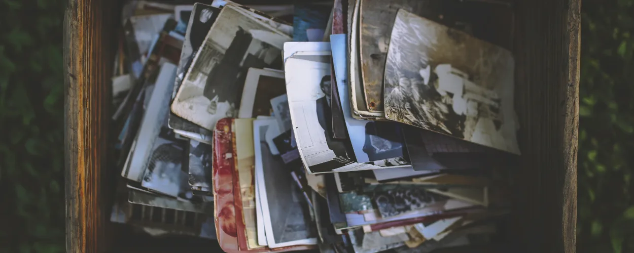 A top-down view of an open wooden crate or box overflowing with a disorganized collection of vintage analog photographs. The stack includes various black and white portraits and snapshots from different eras, showing signs of aging and wear. The box is placed outdoors, surrounded by green foliage, s. How to Price Portrait Print Packages Without Guessing