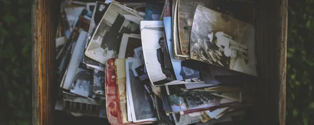 A top-down view of an open wooden crate or box overflowing with a disorganized collection of vintage analog photographs. The stack includes various black and white portraits and snapshots from different eras, showing signs of aging and wear. The box is placed outdoors, surrounded by green foliage, s. How to Price Portrait Print Packages Without Guessing